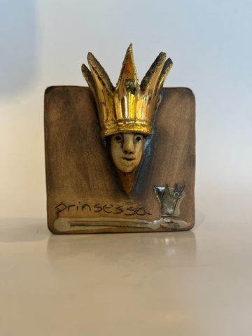 Liten keramikk klosse som viser bilde av en prinsesse med gull krone belagt med gull blader med teksten risset inn: prisessa. Ansiktet og kronen er uthevet. Figuren egnet seg til å henge opp på vegg og et et kunststykke i ethvert rom.